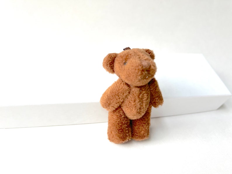 May include: A small brown teddy bear with a soft, plush texture. The bear is standing on a white surface.