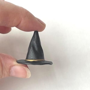 May include: Miniature black witch hat with a gold band around the brim. The hat is held between two fingers against a white background. The hat is a small, handcrafted item.