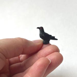 May include: A miniature black bird figurine with red eyes. The small bird is perched upright, with detailed feather textures. The figurine is held in a person's hand, showcasing its small size. The bird is a decorative item.