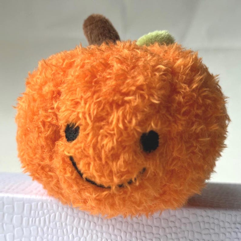 May include: A plush orange pumpkin with a smiling face, black eyes, and a stitched mouth. It has a brown stem and a small green leaf. The pumpkin is a soft, round shape, perfect for fall decor.