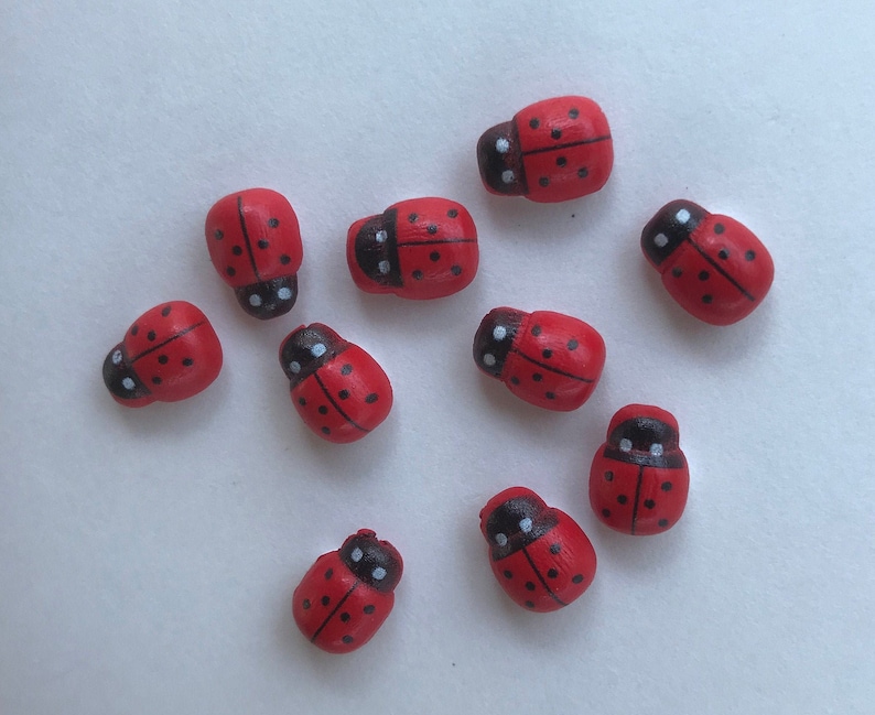 May include: A collection of ten small, rounded wooden beads shaped like ladybugs. Each bead is painted bright red with a black head, a thin black line down the center of the back, and several small black dots scattered across the red surface.