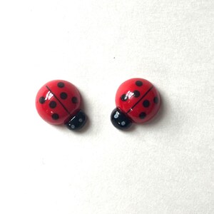 May include: A pair of small, rounded ladybug-shaped cabochons featuring a glossy red finish with black spots and a black head section. Each piece has a smooth, domed surface and a distinct line down the center of the red wings, mimicking a beetle.