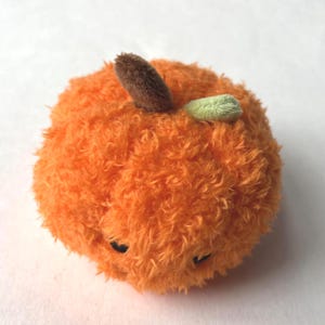May include: A plush orange fruit-shaped toy with a fuzzy texture. It has a brown stem and a small green leaf. The toy has small black eyes and is designed to resemble a tangerine or orange.