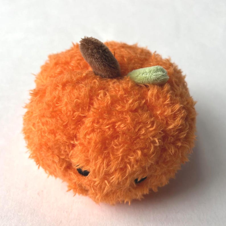 May include: A plush orange fruit-shaped toy with a fuzzy texture. It has a brown stem and a small green leaf. The toy has small black eyes and is designed to resemble a tangerine or orange.
