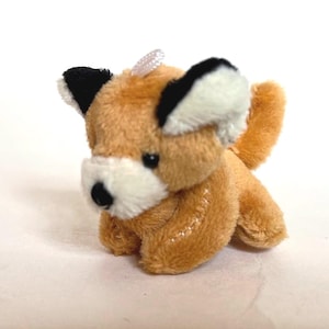 May include: A small, plush fox toy in shades of tan, white, and black. The fox has a white muzzle, black eyes, and black-tipped ears. A small, white ribbon is attached to the top of its head. The toy is made of soft, fuzzy material.