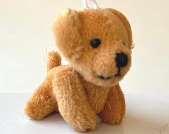 May include: A small, plush golden-brown dog toy with black eyes and nose. The toy has a white loop attached to its head. The dog is in a sitting position, with its front paws extended.
