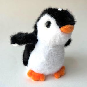 Puede incluir: Un pequeño juguete de peluche de pingüino blanco y negro con patas naranjas y un pico negro. El pingüino está de pie sobre una superficie blanca.