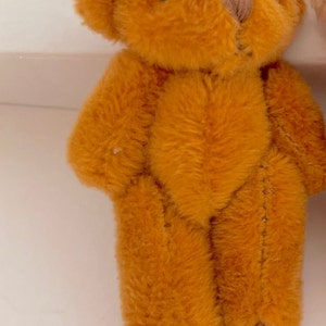 May include: A small, brown teddy bear with a stitched nose and black eyes. The bear is standing on a white surface.