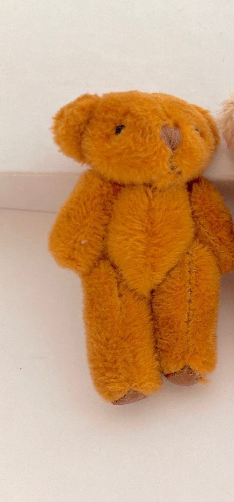 May include: A small, brown teddy bear with a stitched nose and black eyes. The bear is standing on a white surface.