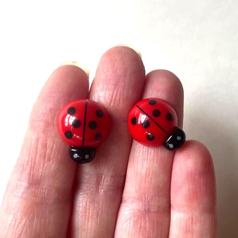 May include: A pair of ladybug-shaped stud earrings featuring a bright red domed surface with distinct black spots and a small black head section. The earrings have a smooth, glossy finish and are designed with a rounded, insect-inspired aesthetic.