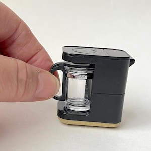 May include: A black miniature coffee maker with a clear glass carafe and a black handle. The coffee maker is on a beige base.