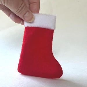 May include: A small red felt Christmas stocking featuring a soft white plush cuff at the top. The stocking has a simple, classic shape with a rounded toe and a straight opening, crafted from a vibrant red fabric with a fuzzy texture.