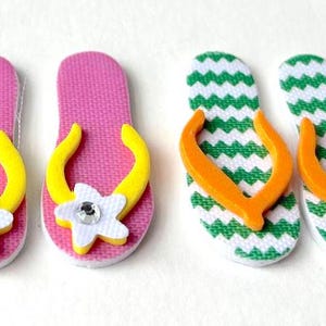 May include: Four pairs of flip-flop style sandals. Two pairs are pink with yellow straps and white flowers with rhinestones. Two pairs are green and white chevron print with orange straps.
