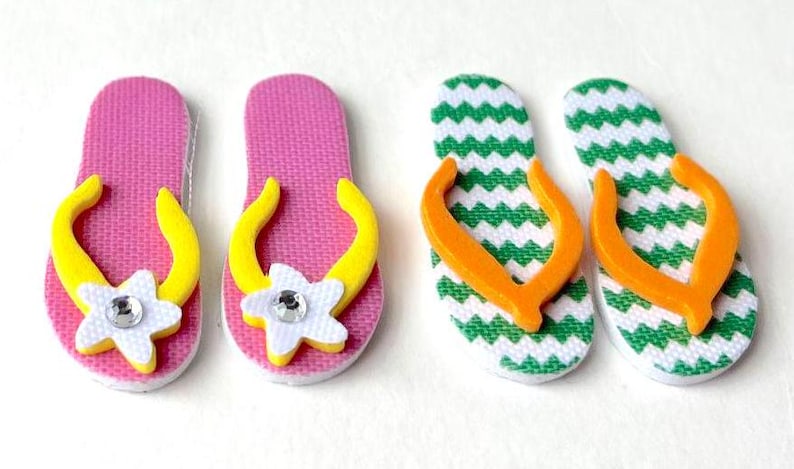 May include: Four pairs of flip-flop style sandals. Two pairs are pink with yellow straps and white flowers with rhinestones. Two pairs are green and white chevron print with orange straps.