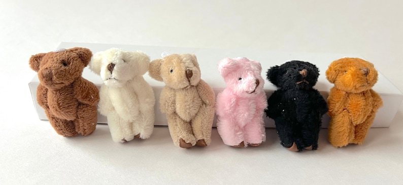 May include: Seven small plush teddy bears in various colours, including brown, white, beige, pink, black, and orange. The bears are all sitting upright with their paws in front of them.