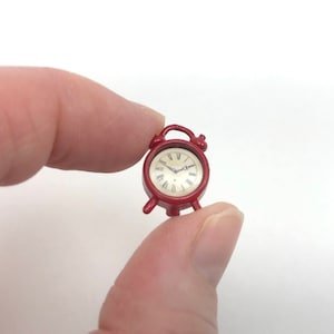 Puede incluir: Un despertador miniatura de color rojo con esfera redonda, fondo crema, números romanos negros y dos manecillas negras finas. El reloj cuenta con una pequeña asa en la parte superior y dos patas cortas en la base, diseñado para exhibición.