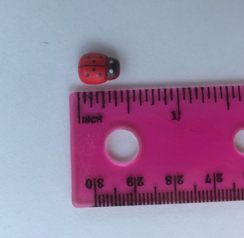 May include: A small, red, dome-shaped ladybug bead with black spots and a dark head, positioned next to a pink plastic ruler. The ruler shows the ladybug bead measures approximately 0.375 inches in length along the imperial scale markings.