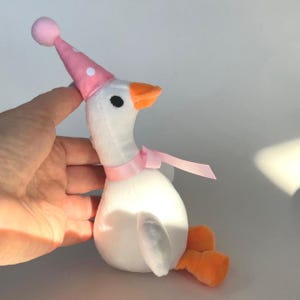 May include: A plush white goose toy with an orange beak and feet, wearing a pink party hat with white polka dots and a pink ribbon. The toy is designed to look like a goose.