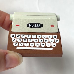Puede incluir: Una máquina de escribir en miniatura con una base marrón y una parte superior color crema. El teclado presenta teclas blancas con letras y símbolos negros. El texto "No.188'" está impreso en la parte superior. Hay detalles en rojo.