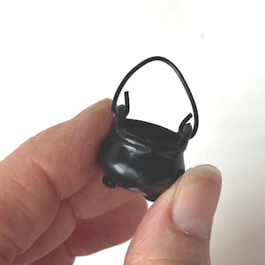 May include: A small, black cauldron with a curved handle. The miniature pot has three small feet and a rounded body. The cauldron is held in a person's hand against a white background. The pot is made of a shiny material.