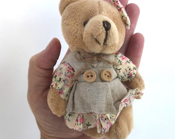 May include: A small, light brown plush teddy bear featuring a floral-patterned fabric with pink and green accents on the sleeves and skirt. The bear wears a beige textured vest with two wooden buttons and a matching floral bow on its head.