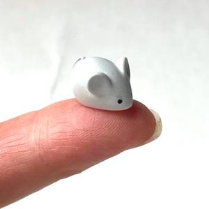 Puede incluir: Una pequeña figura de ratón blanco con ojos negros y orejas redondeadas. El ratón en miniatura está colocado sobre un dedo, mostrando su pequeño tamaño. La figura es de un solo color y tiene una textura suave.