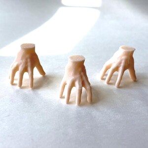 May include: Three small, pale pink, disembodied hands are arranged on a white surface. The hands are posed with fingers slightly curved, and the wrists are cut off. The hands appear to be made of a smooth material.