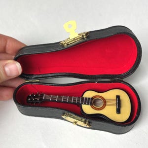 May include: Miniature acoustic guitar in a black case with a red velvet interior. The guitar has a light wood body, a dark fretboard, and a sound hole with a circular design. The case is open, revealing the instrument.