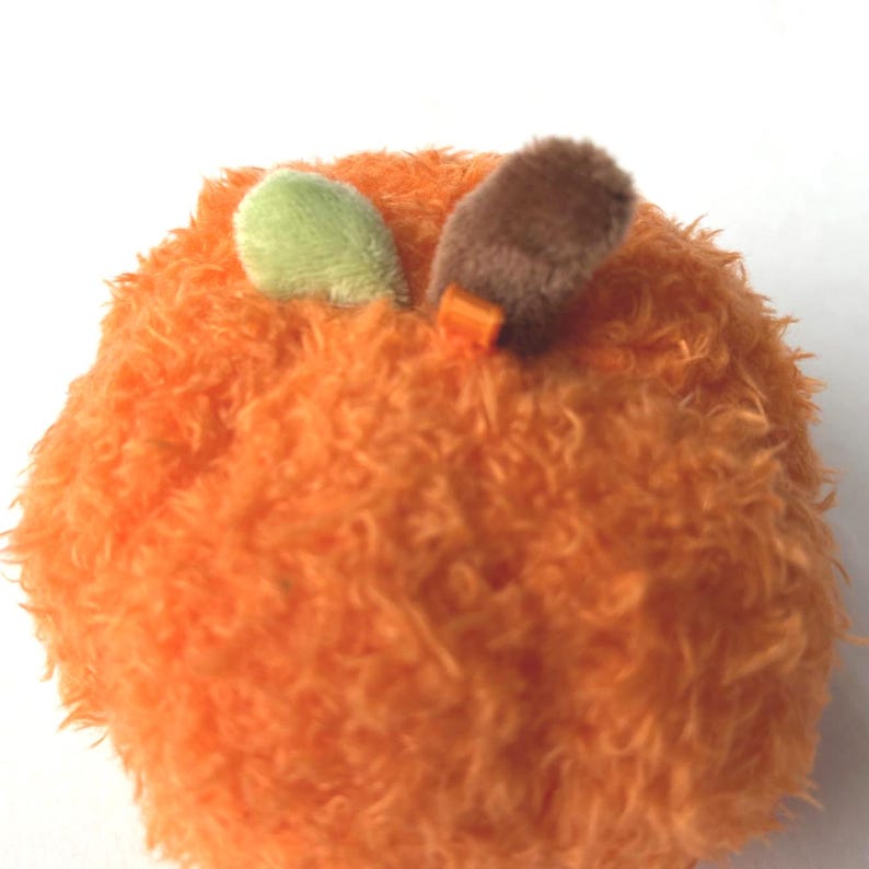May include: A plush orange toy shaped like a fruit, possibly an apple or a pumpkin. It has two small, green and brown leaf-like accents on top, with a small orange tag. The toy has a fuzzy texture.