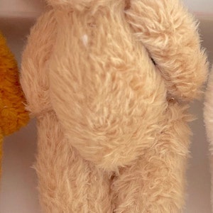 May include: A small, light brown teddy bear with a fluffy texture. The bear is standing on a white surface.