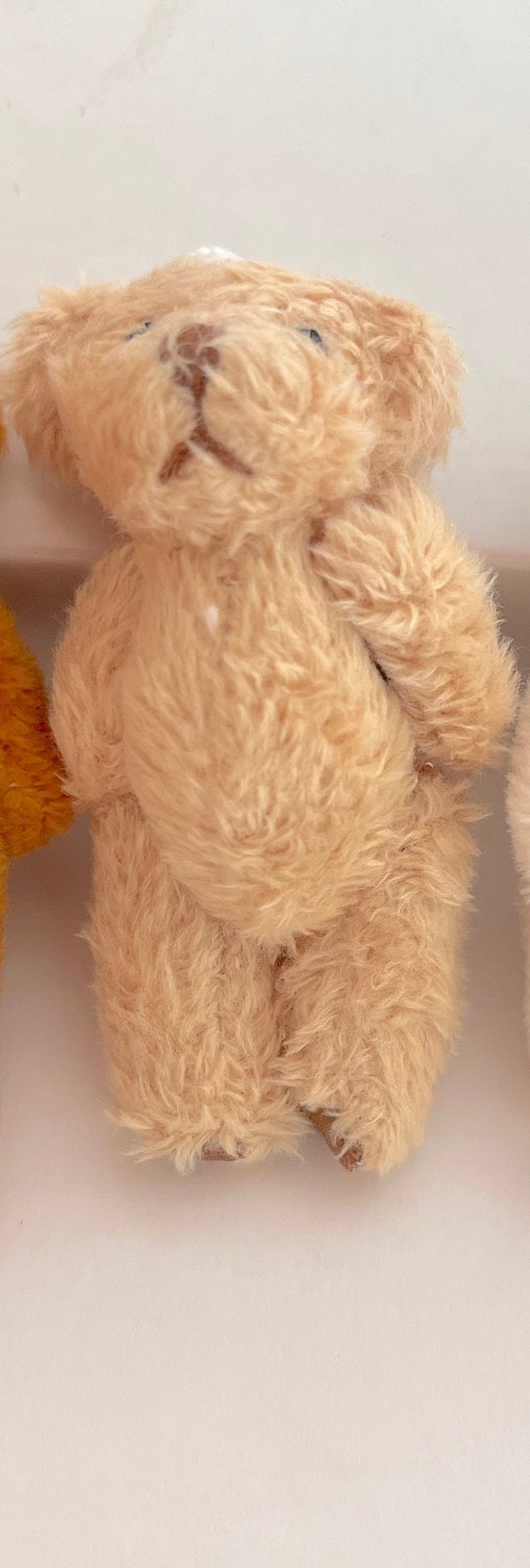 May include: A small, light brown teddy bear with a fluffy texture. The bear is standing on a white surface.