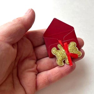 May include: A small red envelope with a gold glitter teddy bear attached with a red ribbon. The envelope is open, revealing a small space inside. The teddy bear is a decorative element.