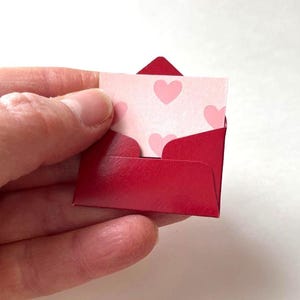 Op de afbeelding: Een kleine rode envelop met een bijpassende rode flap, met een kleine kaart. De kaart is lichtroze met een patroon van roze harten. De envelop wordt in een hand gehouden.