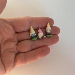 Può includere: Tre gnomi da giardino in miniatura con cappelli gialli, rosa e bianchi. Gli gnomi hanno abiti colorati e sono in piedi su basi verdi. Gli gnomi sono piccoli, alti circa 2,5 cm, e sono tenuti in una mano.