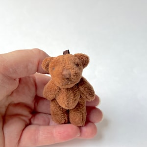 May include: A small brown teddy bear with a brown nose and black eyes. The bear is standing on a person's hand.