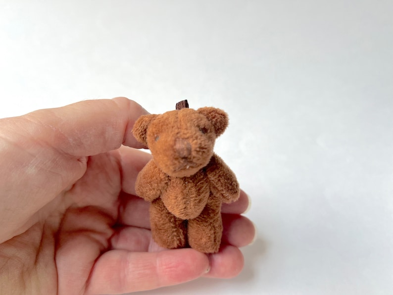 May include: A small brown teddy bear with a brown nose and black eyes. The bear is standing on a person's hand.