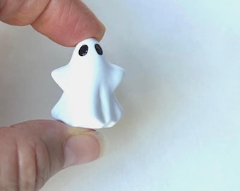 May include: A small, white, ghost-shaped figurine with a smooth, matte finish. The ghost features two simple, black, oval-shaped eyes positioned near the top of its rounded head, with draped, flowing edges that mimic a sheet-like appearance.