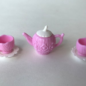 May include: A pink tea set with a teapot, two cups, and saucers. The teapot has a white lid and a decorative design. The cups and saucers are also decorated with a similar design.