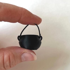 May include: A small, black cauldron with a curved handle. The miniature pot has two small handles on either side and is held between two fingers. The cauldron is likely a decorative item or part of a miniature set.