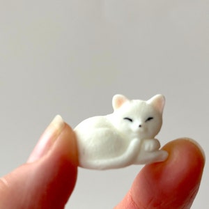 May include: A small white ceramic figurine of a cat sleeping on its side. The cat has closed eyes and a small, curled tail.