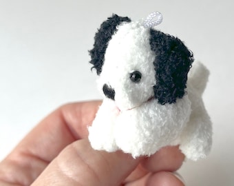May include: A small white and black plush dog toy with a white loop on top of its head. The dog is sitting on a hand.