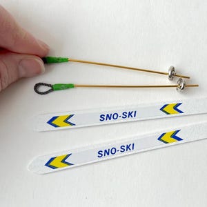 May include: Two white skis with yellow and blue arrows and the text "SNO-SKI" printed on them. Two gold ski poles with green handles and silver metal tips.