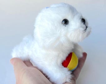 Puede incluir: Un peluche de foca blanca con ojos negros y una pequeña pelota roja, amarilla y azul. La foca está hecha de material suave y esponjoso y tiene una expresión amigable. El juguete está diseñado para ser tierno y atractivo.