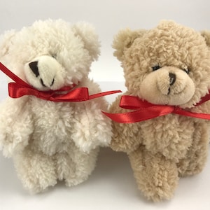 May include: Two plush teddy bears with red ribbon bows. The bear on the left is white and the bear on the right is beige.