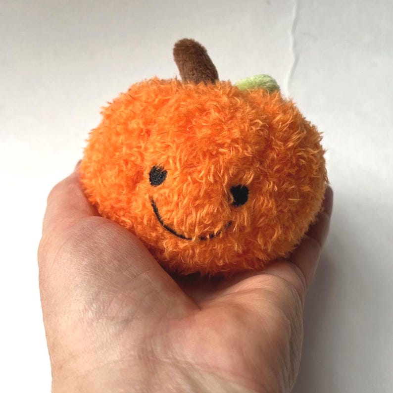 May include: A plush orange pumpkin with a smiling face, brown stem, and green leaf. The pumpkin is round and fluffy, perfect for fall decor or a child's toy.