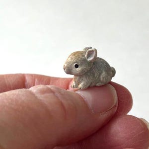 May include: A miniature figurine of a small, light brown bunny with black eyes and detailed fur texture. The bunny is sitting upright, showcasing its small ears and rounded body. The figurine is held on a finger.