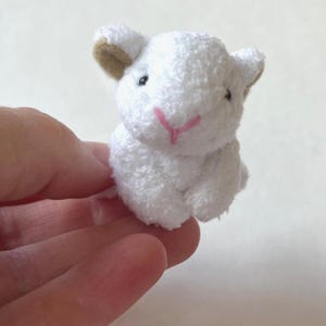 Könnte beinhalten: Ein kleines, weißes Plüschkaninchen mit flauschiger Textur. Das Kaninchen hat braune Ohren, schwarze Augen und eine rosa genähte Nase. Das Spielzeug ist so konzipiert, dass es aufrecht sitzt und seine geringe Größe und sein weiches Aussehen hervorhebt.