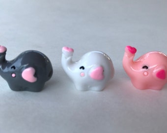 May include: Three small elephant figurines in dark gray, white, and light pink. Each elephant features a glossy finish, a heart-shaped detail on the trunk, a heart-shaped ear, and small painted eyes with a dot for a cheek.