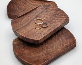 May include: Three rectangular dark brown wooden trays with curved edges stacked in a staggered arrangement. The middle tray holds three simple metal rings, including one gold-toned band and two silver-toned bands, resting on the wood surface.