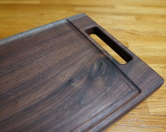 May include: A rectangular, dark brown wooden cutting board with a handle. The board has a recessed area around the edge and a smooth, polished surface. The wood grain is visible, adding to its natural aesthetic.